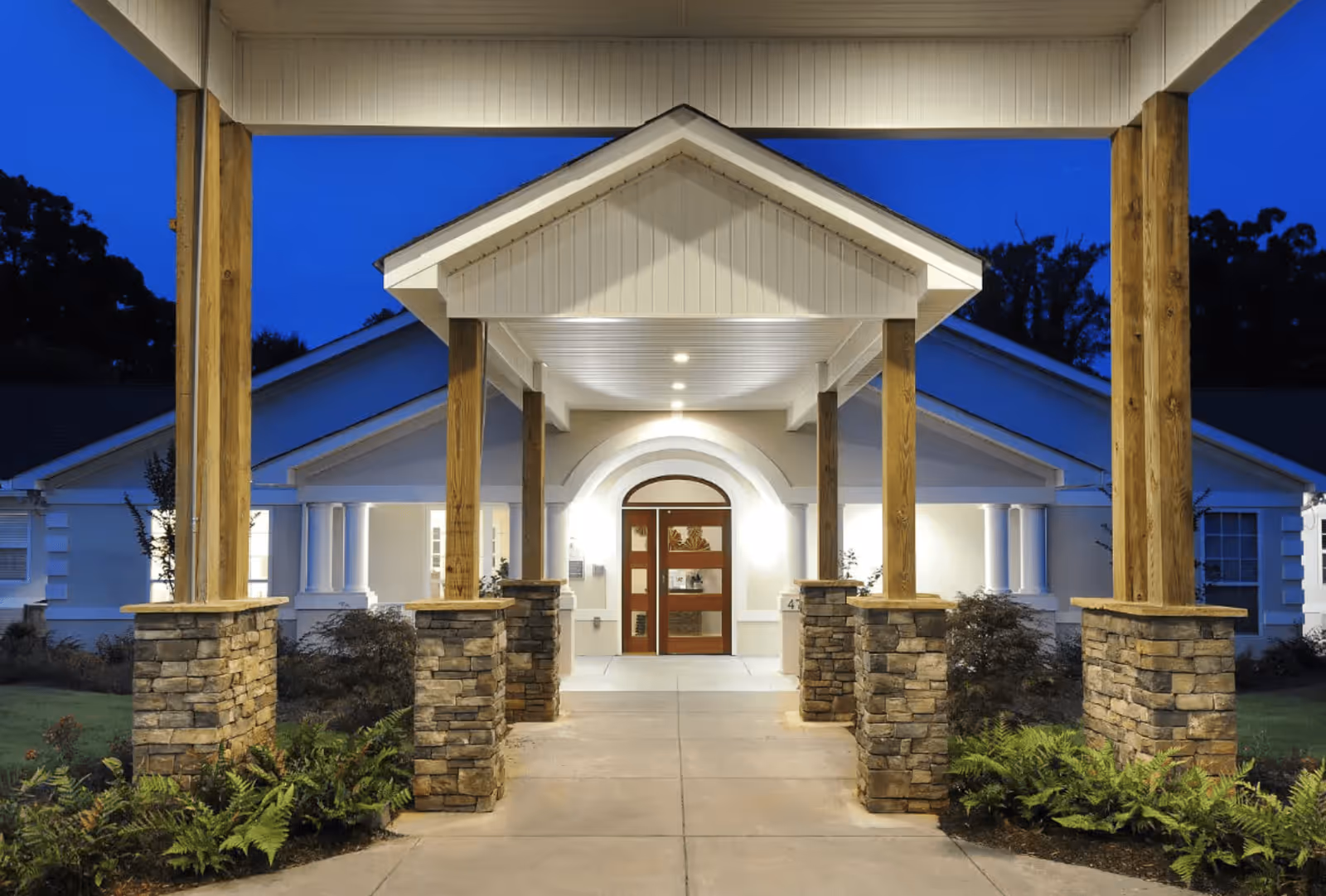 Entrance of a senior living facility at dusk with a covered walkway supported by wooden pillars on stone bases, leading to a well-lit door with an arched frame. The building is white with a pitched roof and surrounded by landscaping including bushes and ferns.