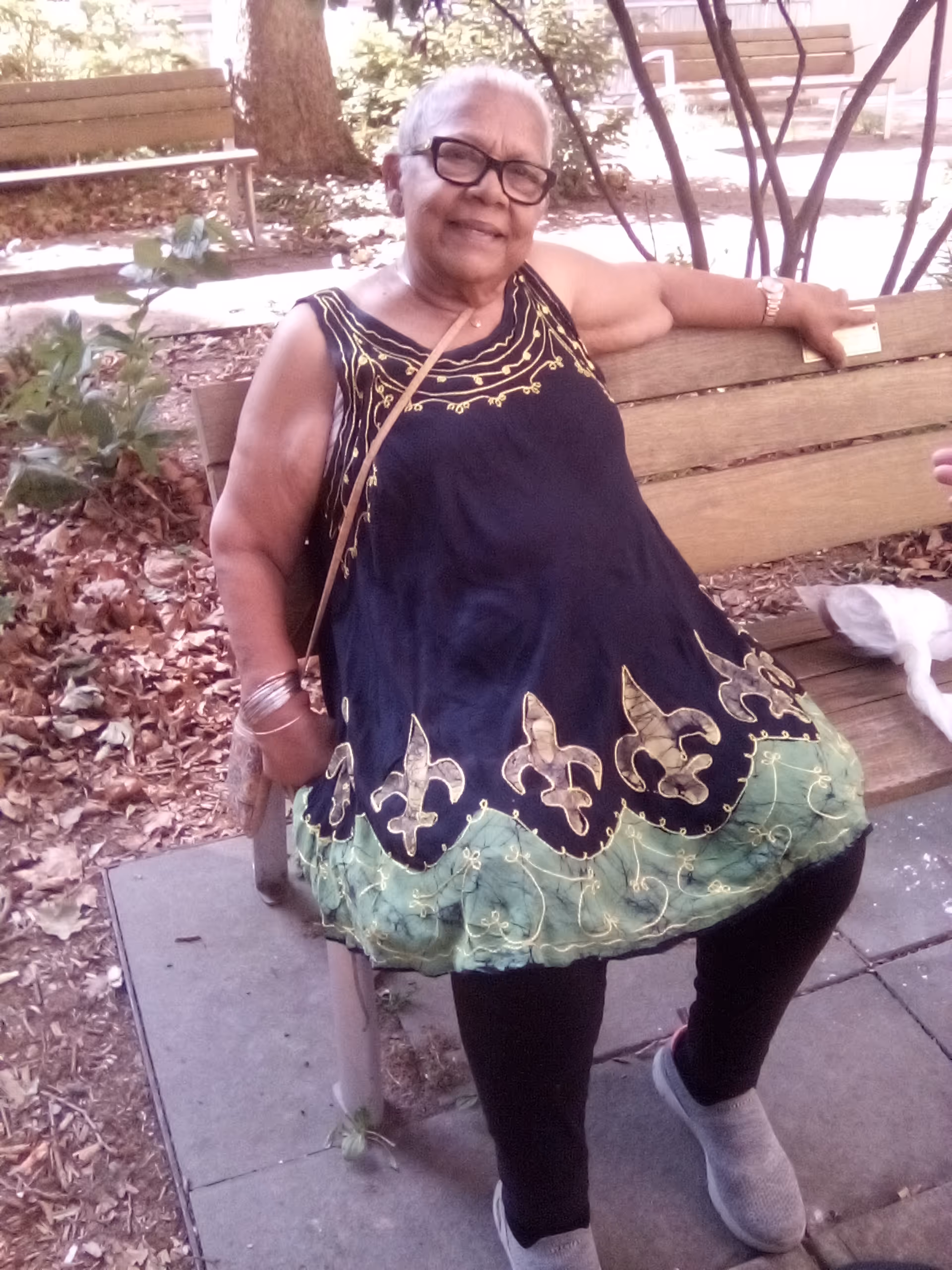 Elderly woman sits on an outdoor park bench, smiling, wearing a dark decorative dress.