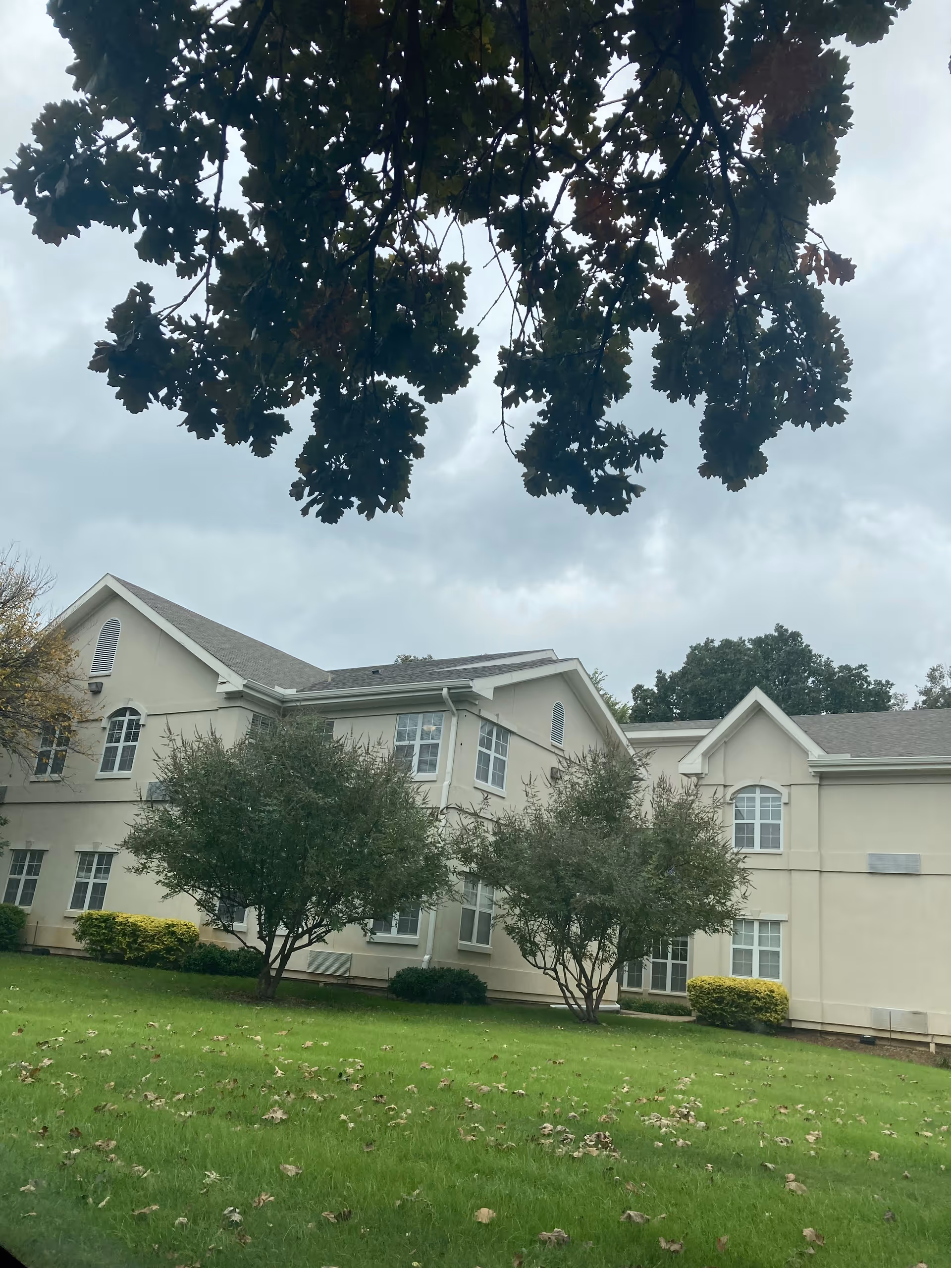 Beige senior living facility exterior with large windows, green lawn, and trees.