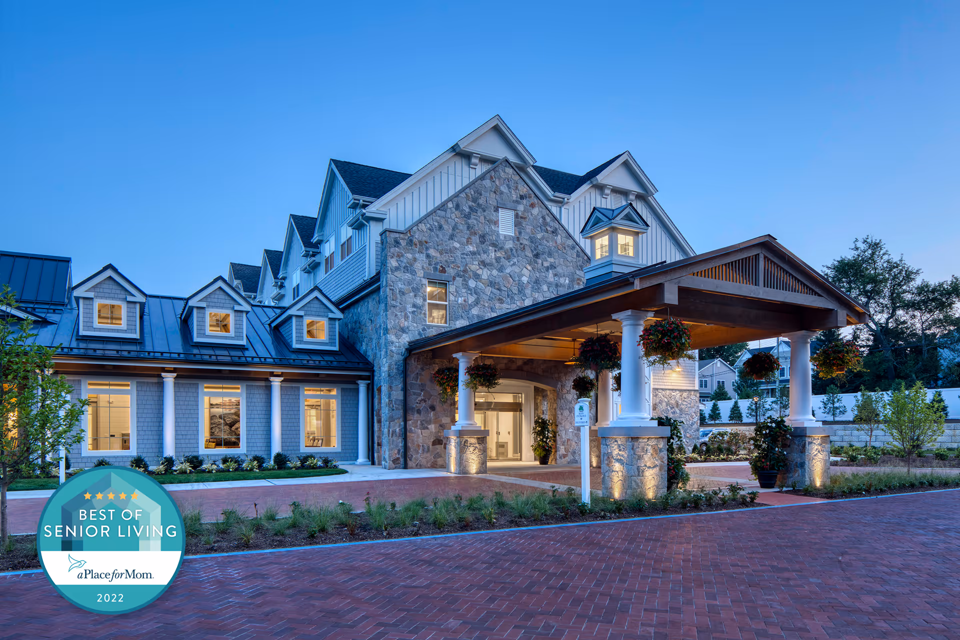 Exterior view of Maplewood at Southport senior living facility at dusk, featuring a large stone and siding building with a covered entrance supported by white columns, hanging flower baskets, and well-maintained landscaping with a brick driveway.