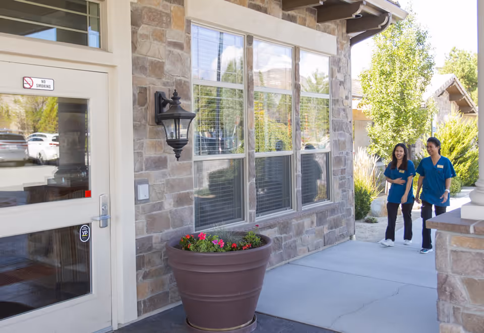Entrance of a stone-faced assisted living building with large windows, a potted flower planter, and two staff in teal scrubs walking on the sidewalk.