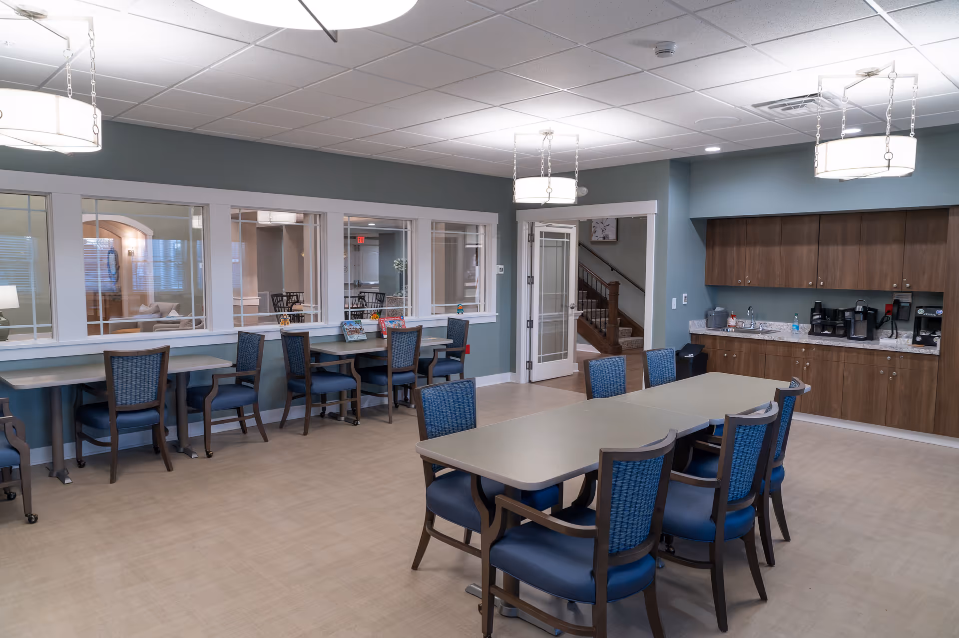 Interior dining area with a long table, blue chairs, and a kitchenette along the wall.