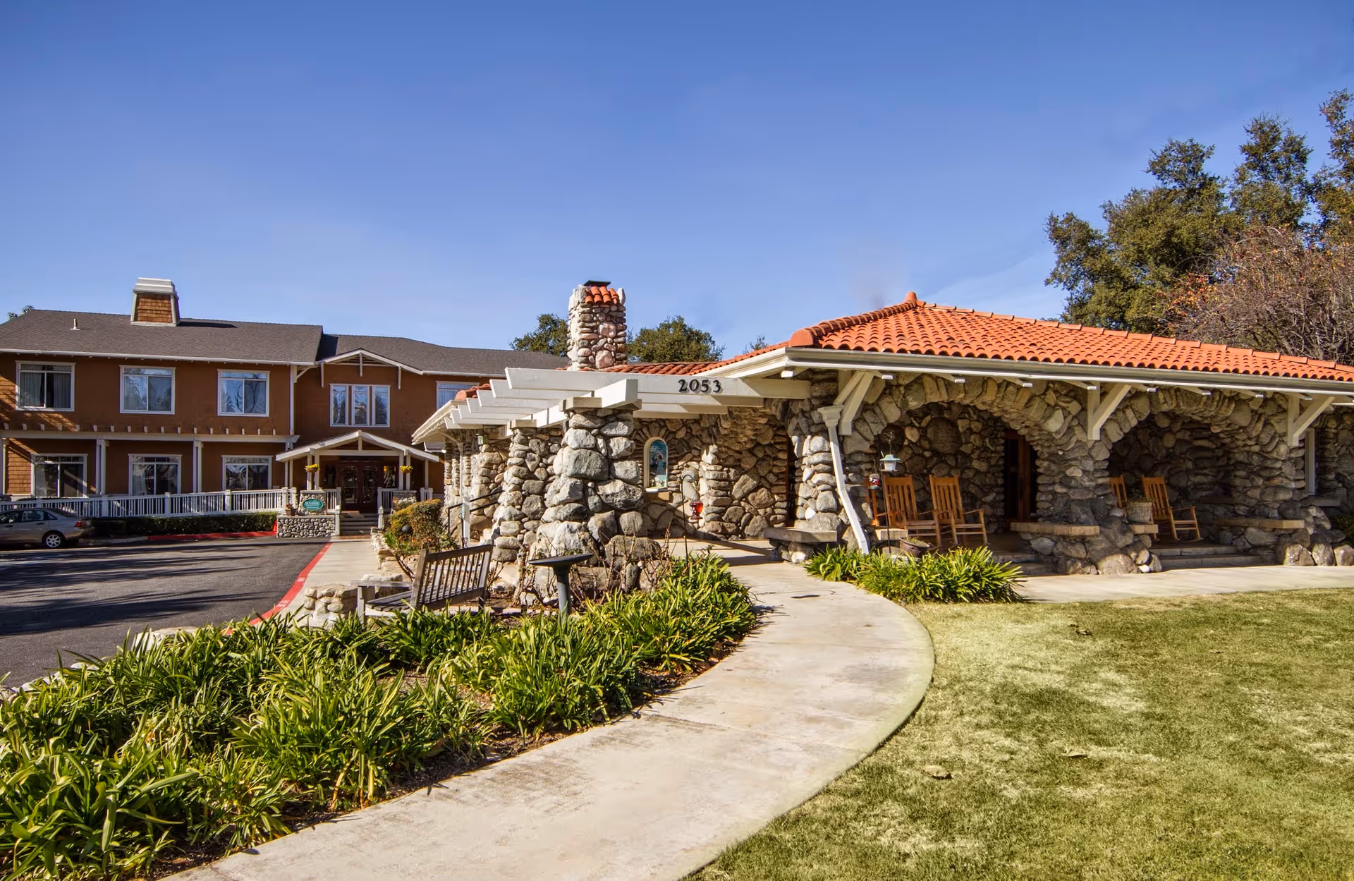 Exterior view of Ivy Park at Claremont senior living facility showing a stone building with a red tiled roof and a pathway leading to the entrance. There are rocking chairs on the porch and greenery along the walkway. The main building with multiple windows is visible in the background under a clear blue sky.