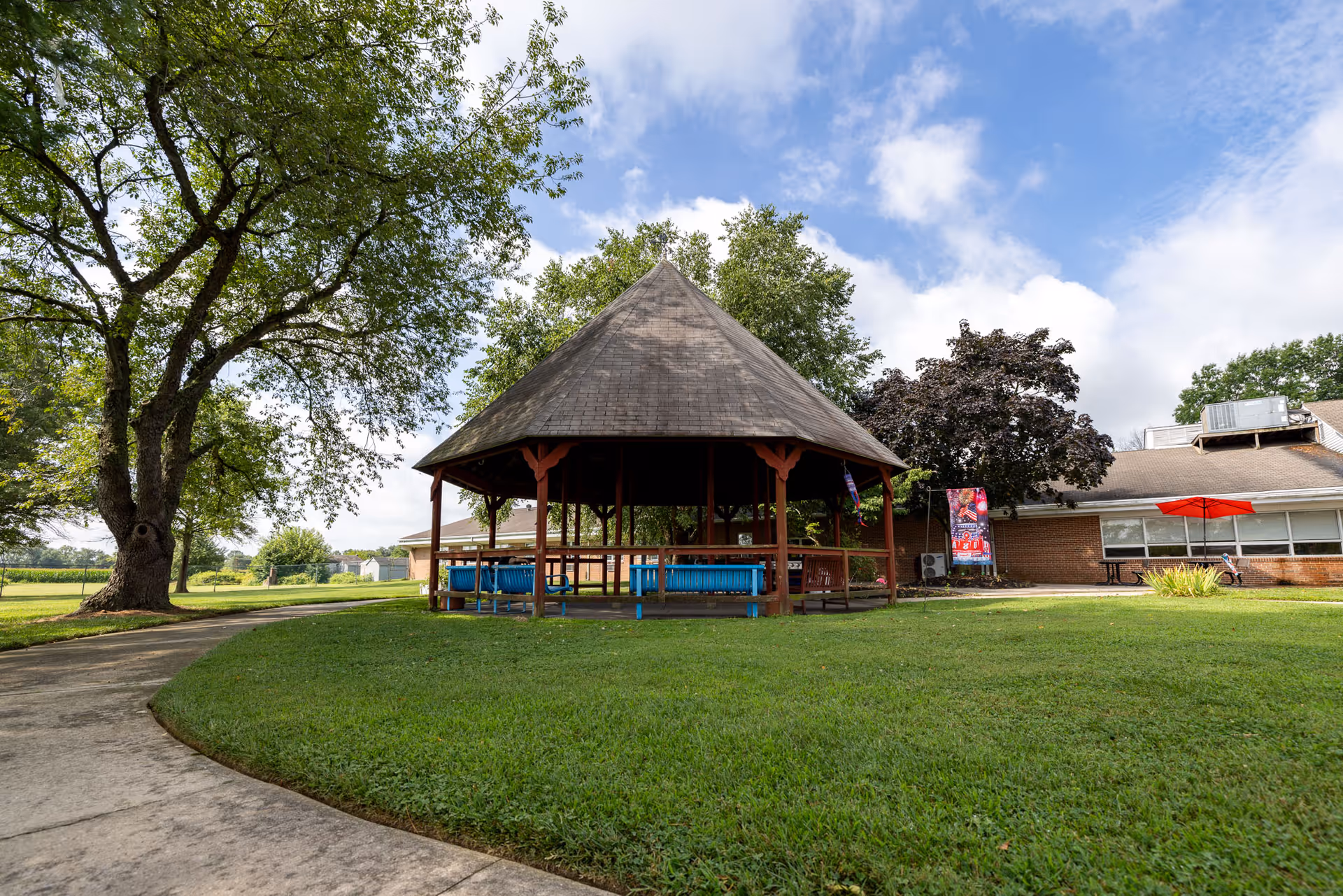 A large outdoor gazebo with a pointed roof surrounded by green grass and trees. There is a curved concrete pathway leading to the gazebo. In the background, there is a brick building with windows and a red umbrella over a picnic table. The sky is partly cloudy with patches of blue.