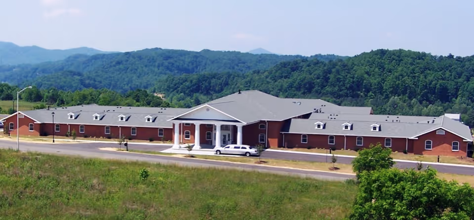 Front view of a long single-story brick assisted-living building with a columned entrance set against wooded hills.