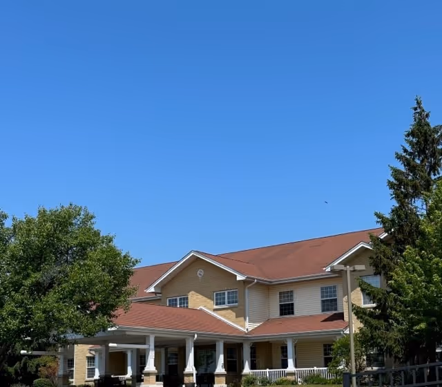 Senior living facility with beige siding, red roof, and a covered front entrance amid trees.