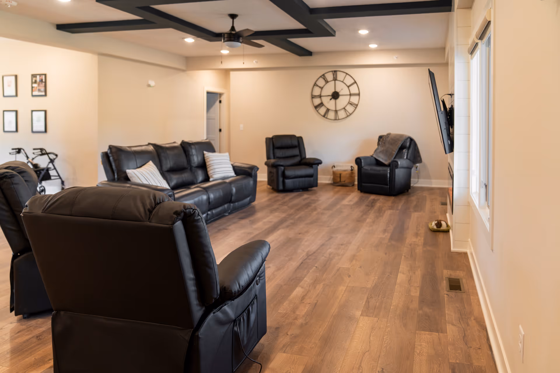 Spacious senior living lounge with multiple black leather recliners, wood floor, and a wall clock.