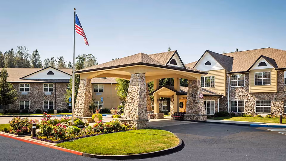 Exterior view of a senior living facility with a covered entrance supported by stone pillars, an American flag on a flagpole, landscaped flower beds, and a two-story building with stone and beige siding under a clear blue sky.