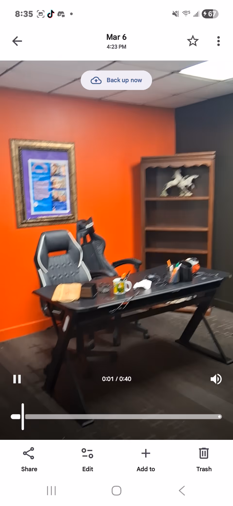 An office-style workstation with a black desk, gaming chair, orange wall, and bookshelf.