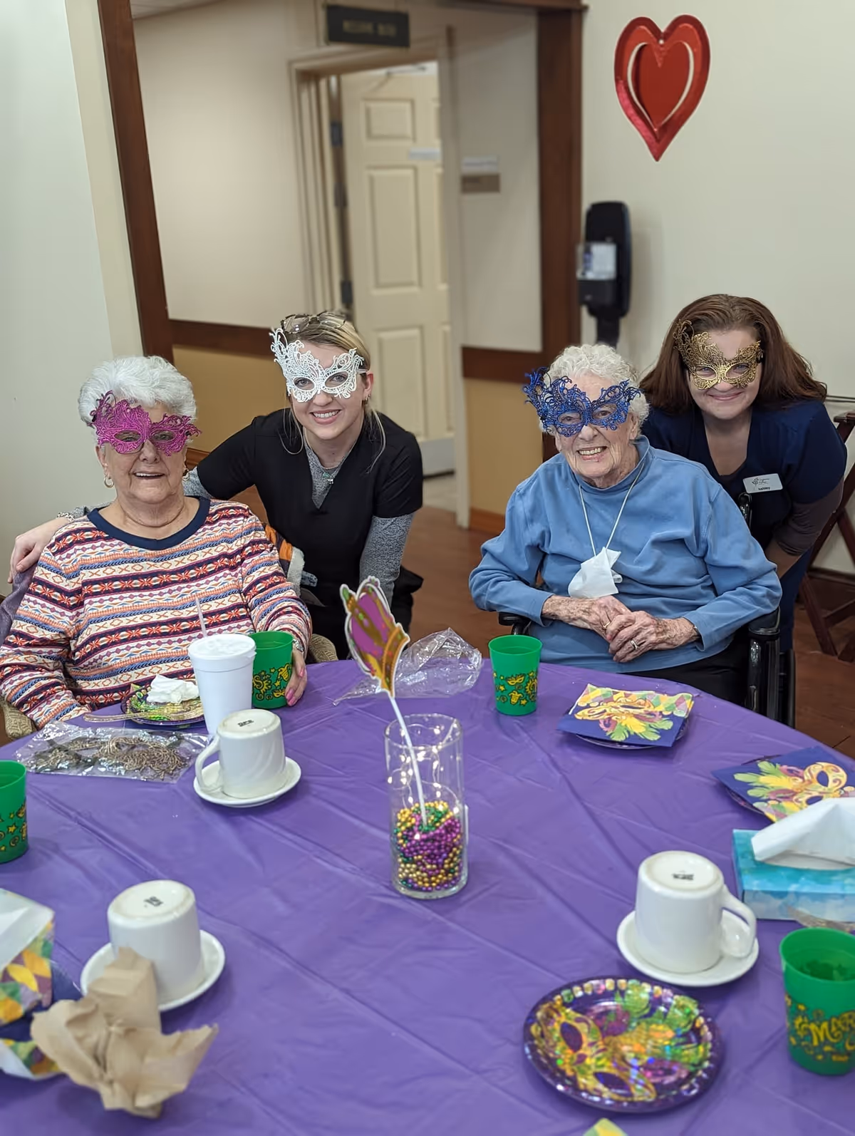 Two elderly women and two caregivers wearing decorative masquerade masks sitting and standing around a table covered with a purple tablecloth. The table has colorful Mardi Gras-themed cups, plates, napkins, and a centerpiece with beads. A red heart decoration hangs on the wall behind them.
