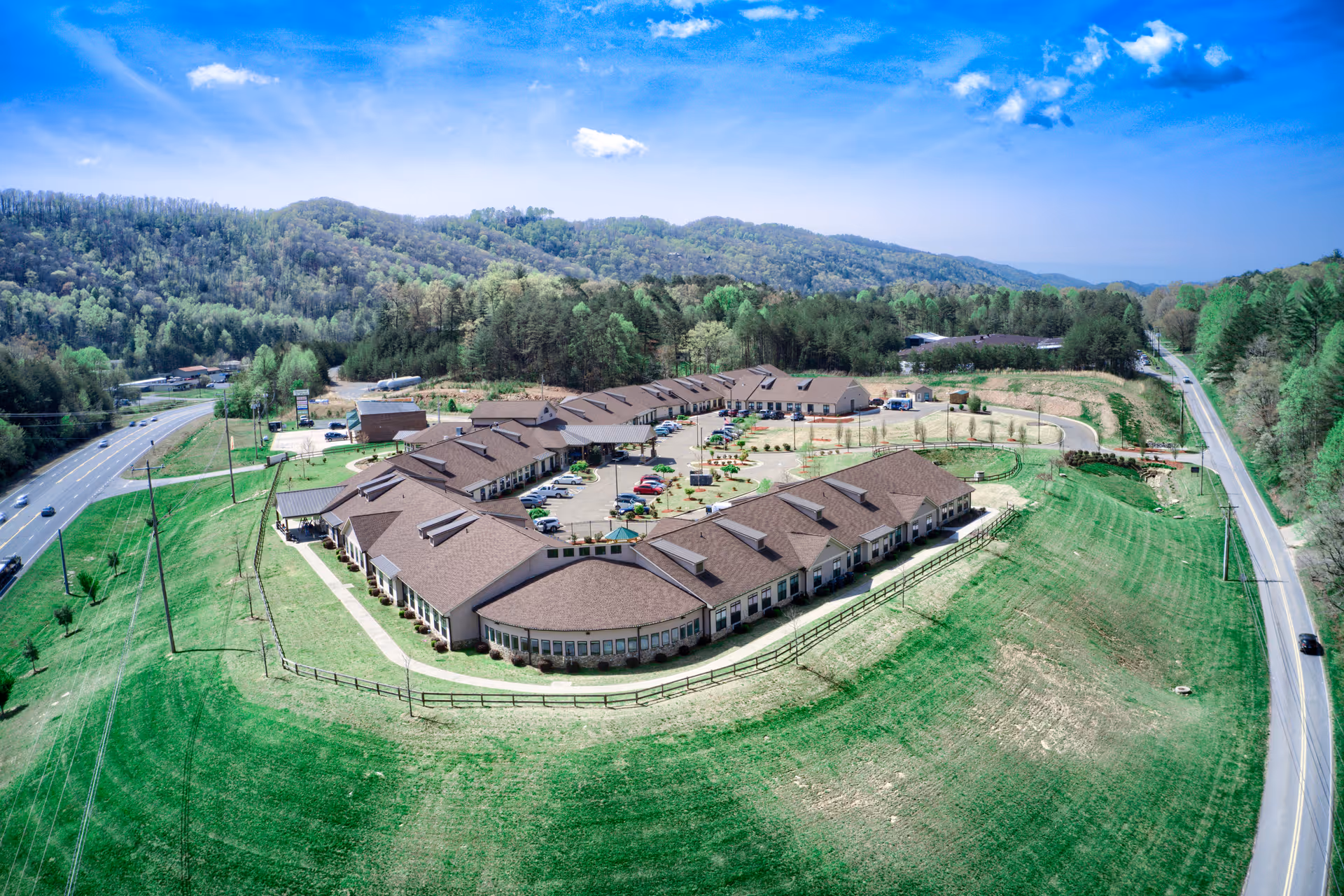 Aerial view of a single-story assisted living complex with connected buildings, parking lot and surrounding lawns set among rolling wooded hills.