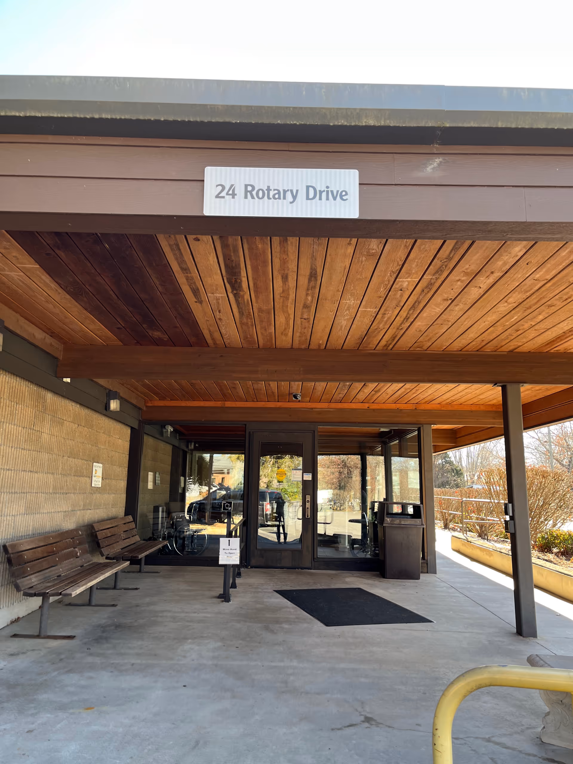 Exterior entrance of a building with glass doors, wooden overhang, and benches outside.