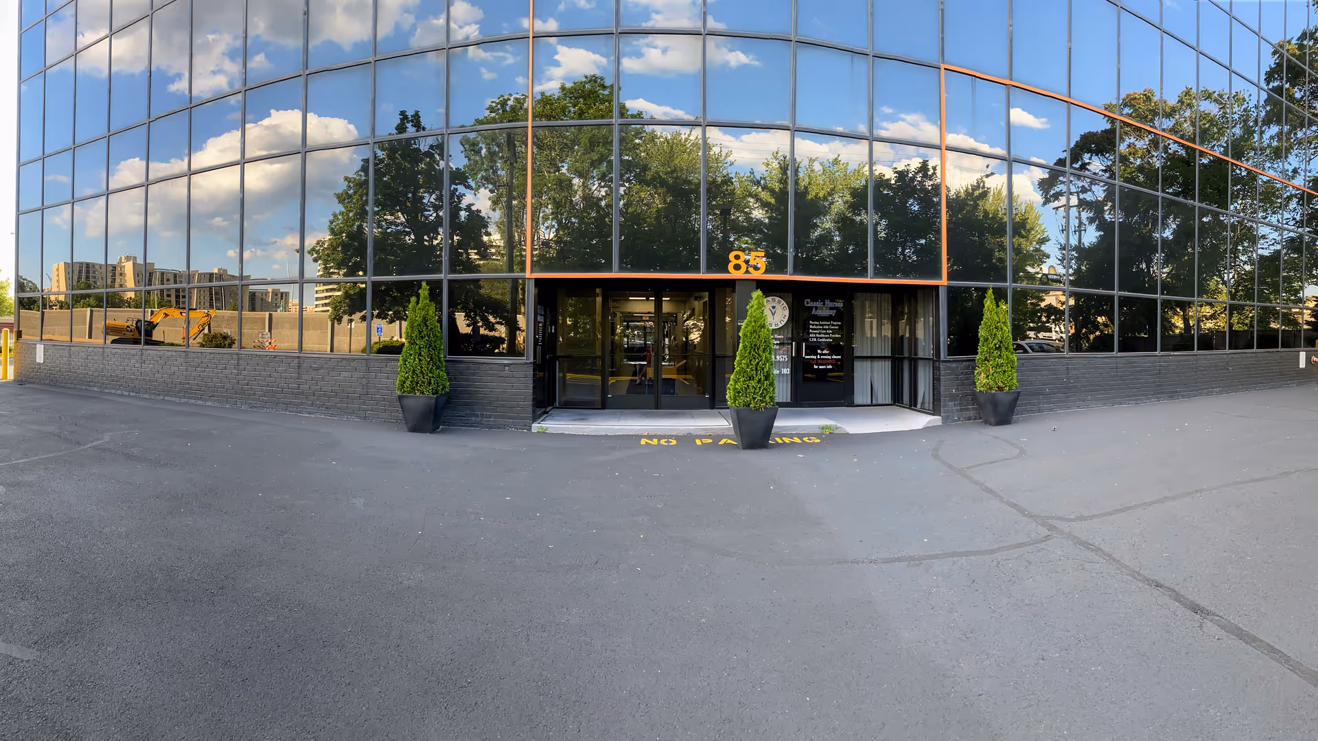 Glass-front facility entrance with orange trim and potted evergreens outside.