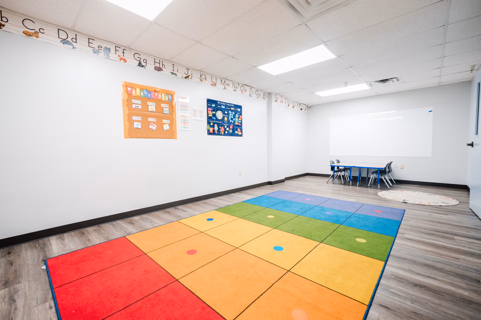 Bright activity room with a colorful square rug, whiteboard, posters, and a small table with chairs.