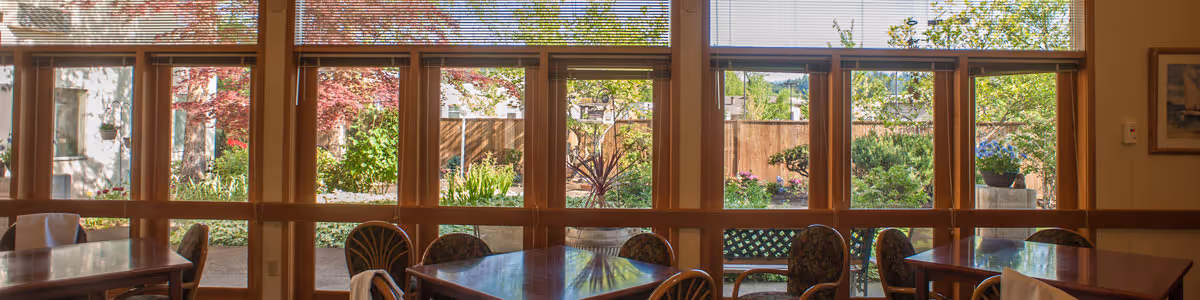 Sunlit dining room with wooden tables and patterned chairs along large windows looking out to a landscaped garden.