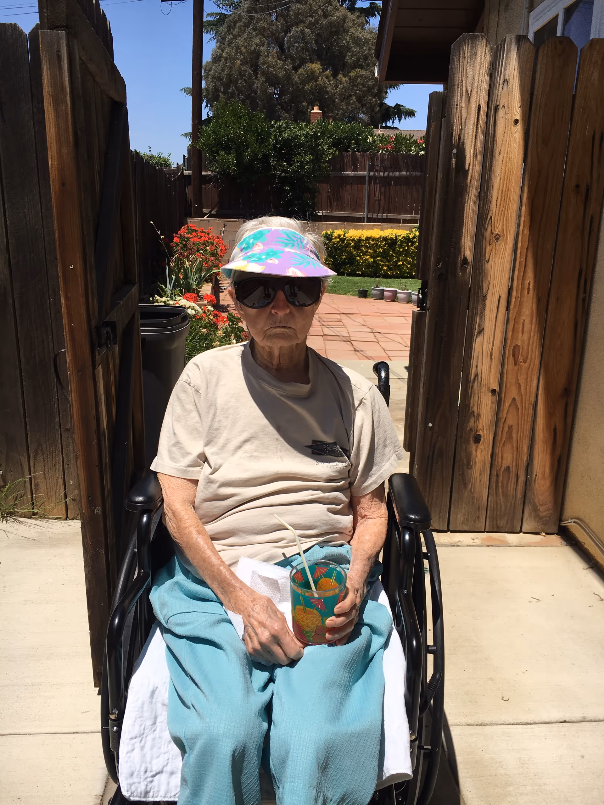 An elderly person sitting in a wheelchair outdoors on a sunny day, wearing a light-colored t-shirt, teal pants, a colorful visor, and large sunglasses, holding a cup with a straw. The background shows a wooden fence, some plants, and a paved patio area.