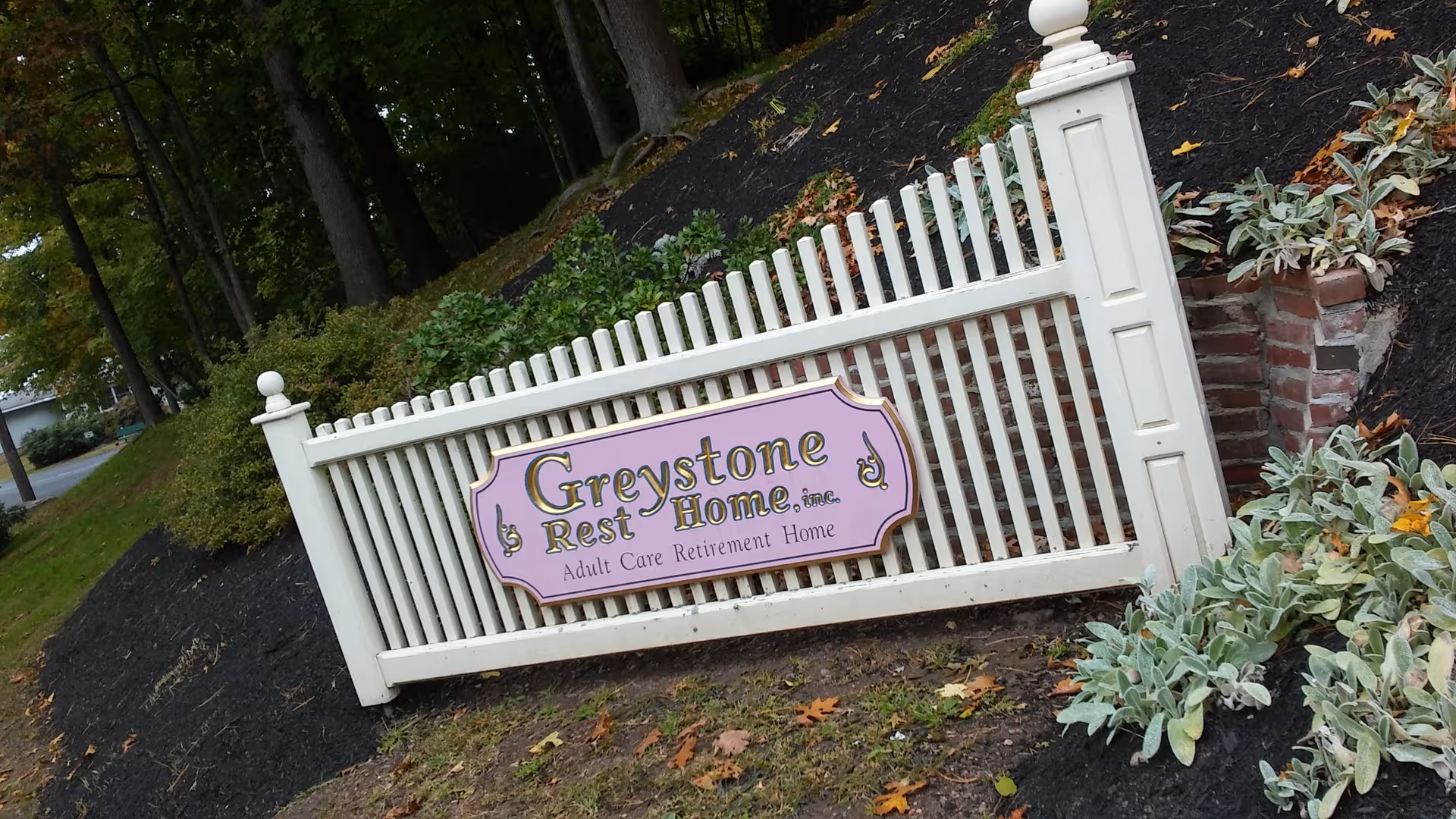 White decorative fence and lavender sign reading Greystone Rest Home at landscaped entrance.