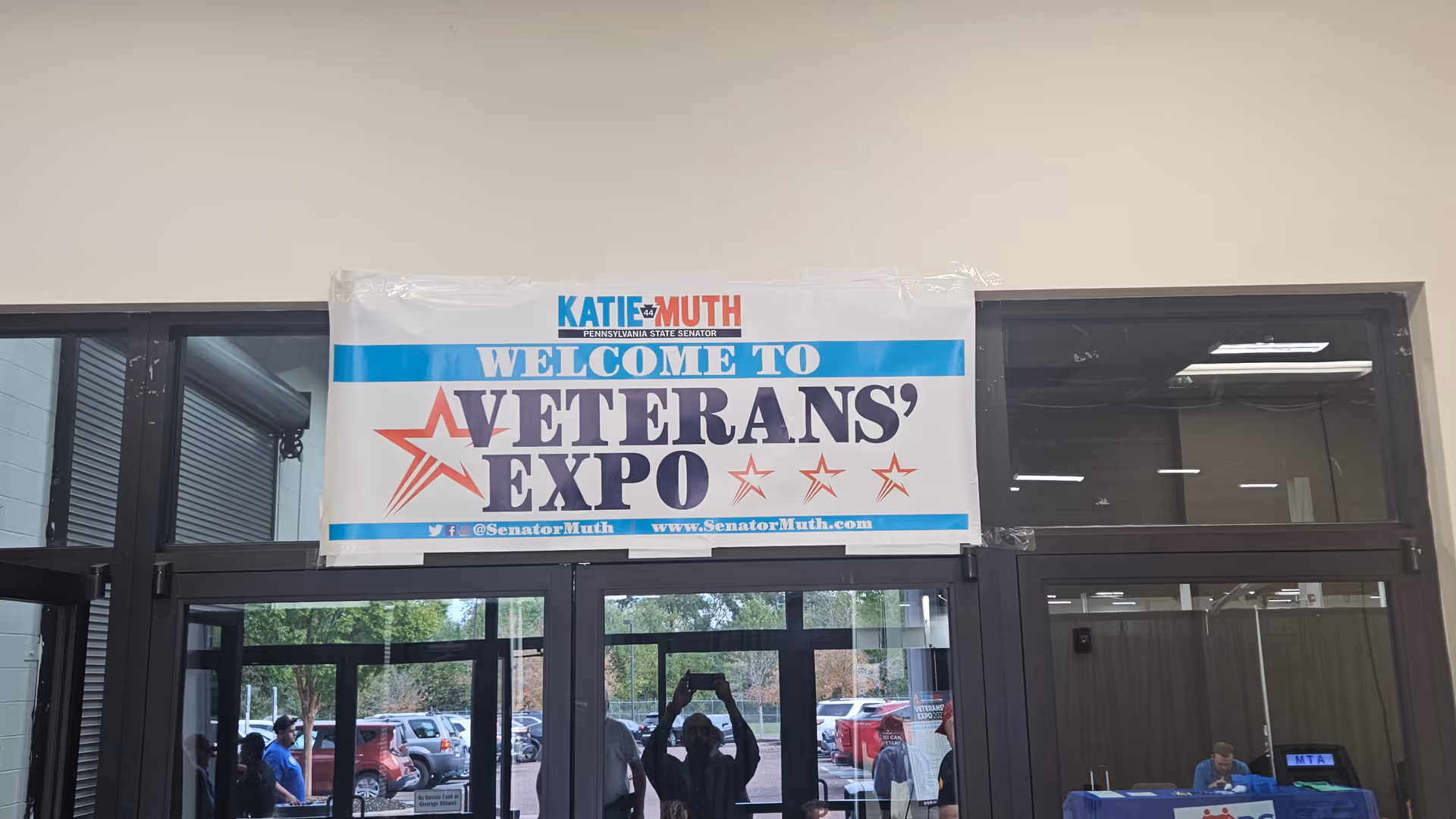 Entrance doors with a Veterans' Expo banner overhead and people outside near parked cars.