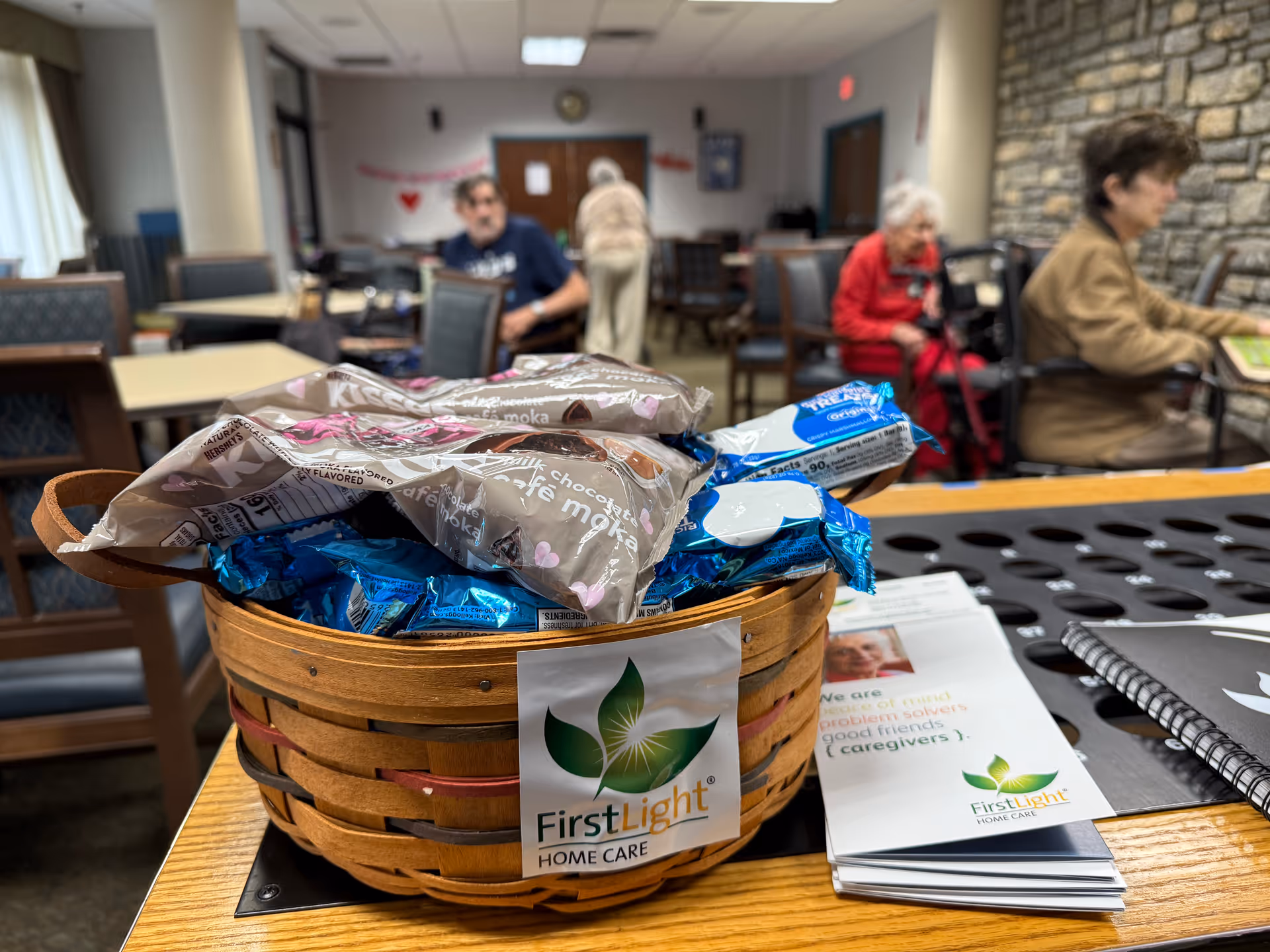 Basket of wrapped snacks labeled FirstLight sits on a wooden table, with residents in a lounge.