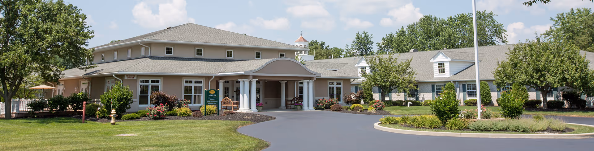 Exterior view of a single-story senior living facility building with a covered entrance, surrounded by well-maintained landscaping including trees, bushes, and flowers under a partly cloudy sky.