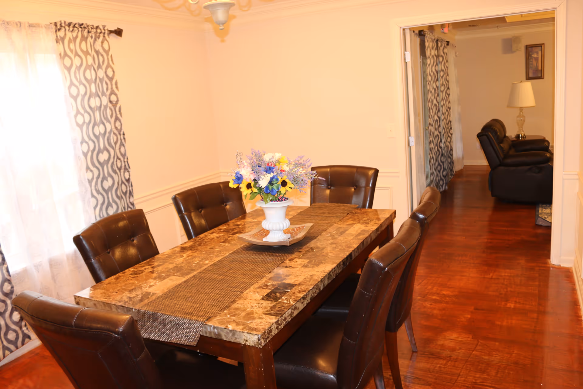 A dining room with a rectangular marble-top table surrounded by six dark brown leather chairs. A vase with colorful artificial flowers is placed in the center of the table on a brown table runner. The room has light-colored walls, patterned curtains on the window, and a wooden floor. In the background, there is a view into a living room area with dark leather recliners and a lamp on a side table.