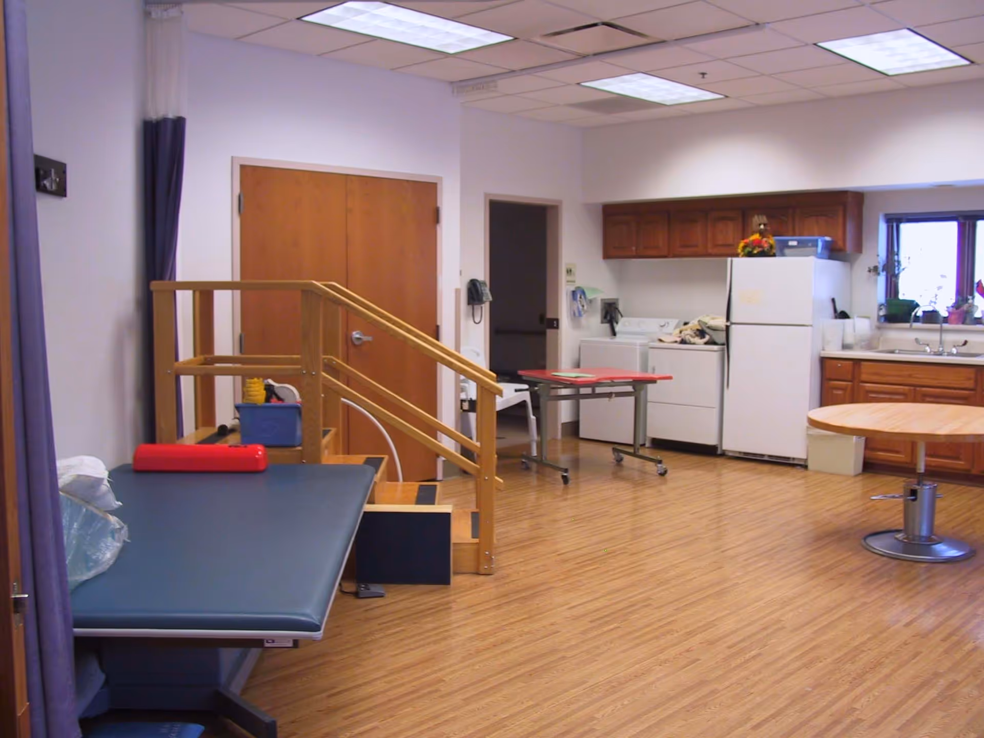 Rehabilitation/therapy room with a treatment table, wooden practice stairs, and a kitchenette area with a refrigerator and sink.