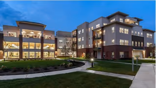 Exterior of a modern, multi-story senior living facility at dusk with landscaped lawn and walkways.