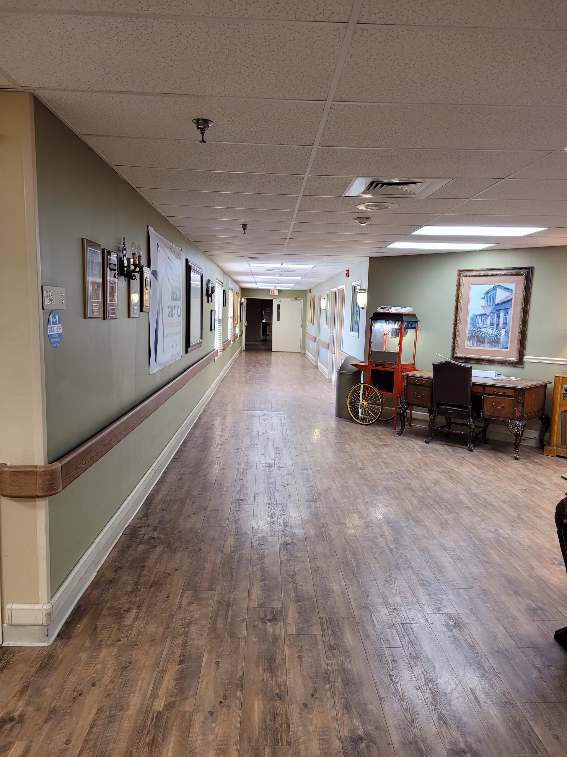 A long wood-floored interior hallway in a senior living facility with handrails, framed art, and a desk with a popcorn cart on the right.