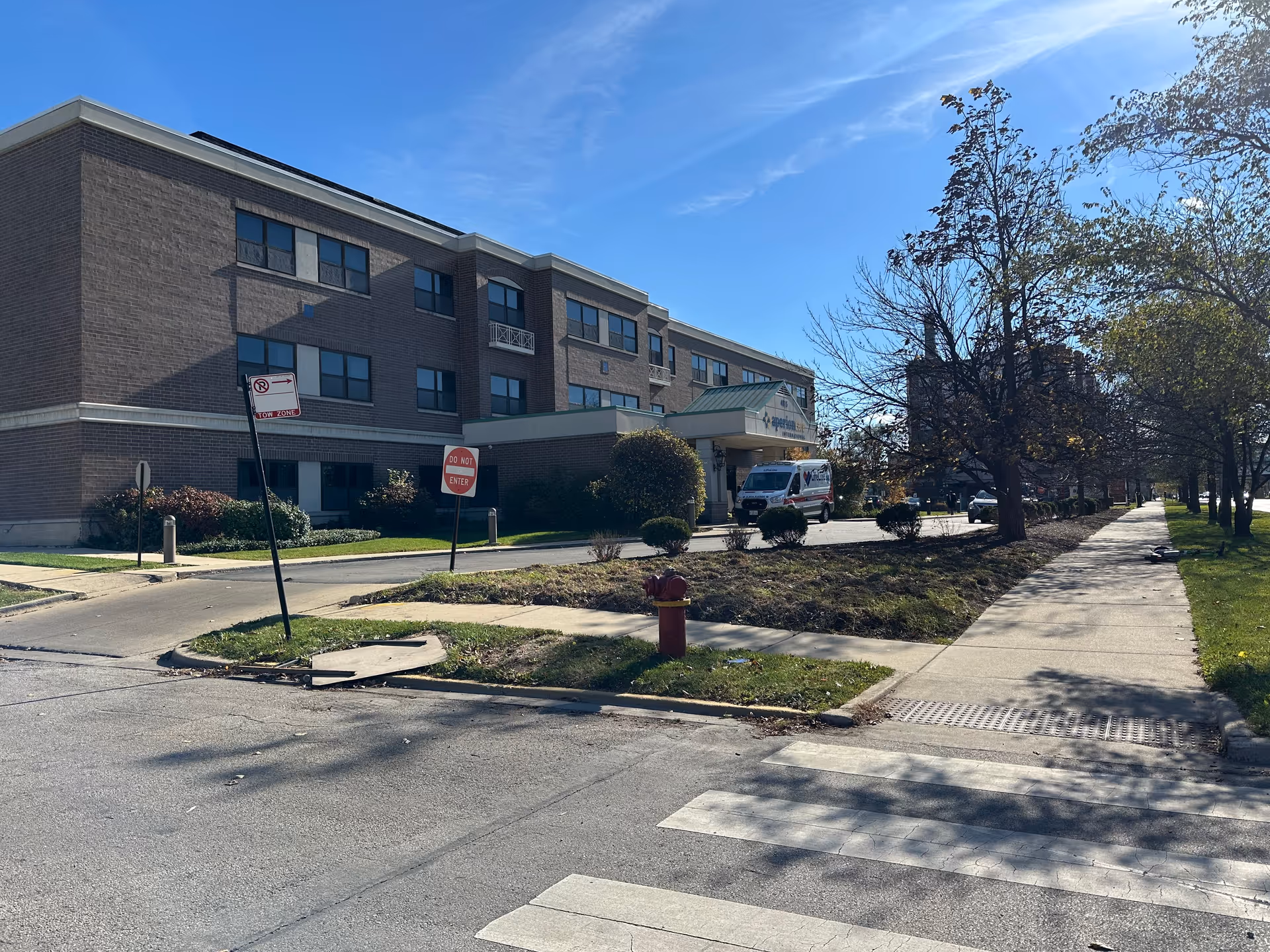 Exterior view of a three-story brick building under a clear blue sky, with a driveway leading to the entrance where an ambulance is parked. There are trees and bushes along the sidewalk and a fire hydrant near the street corner.