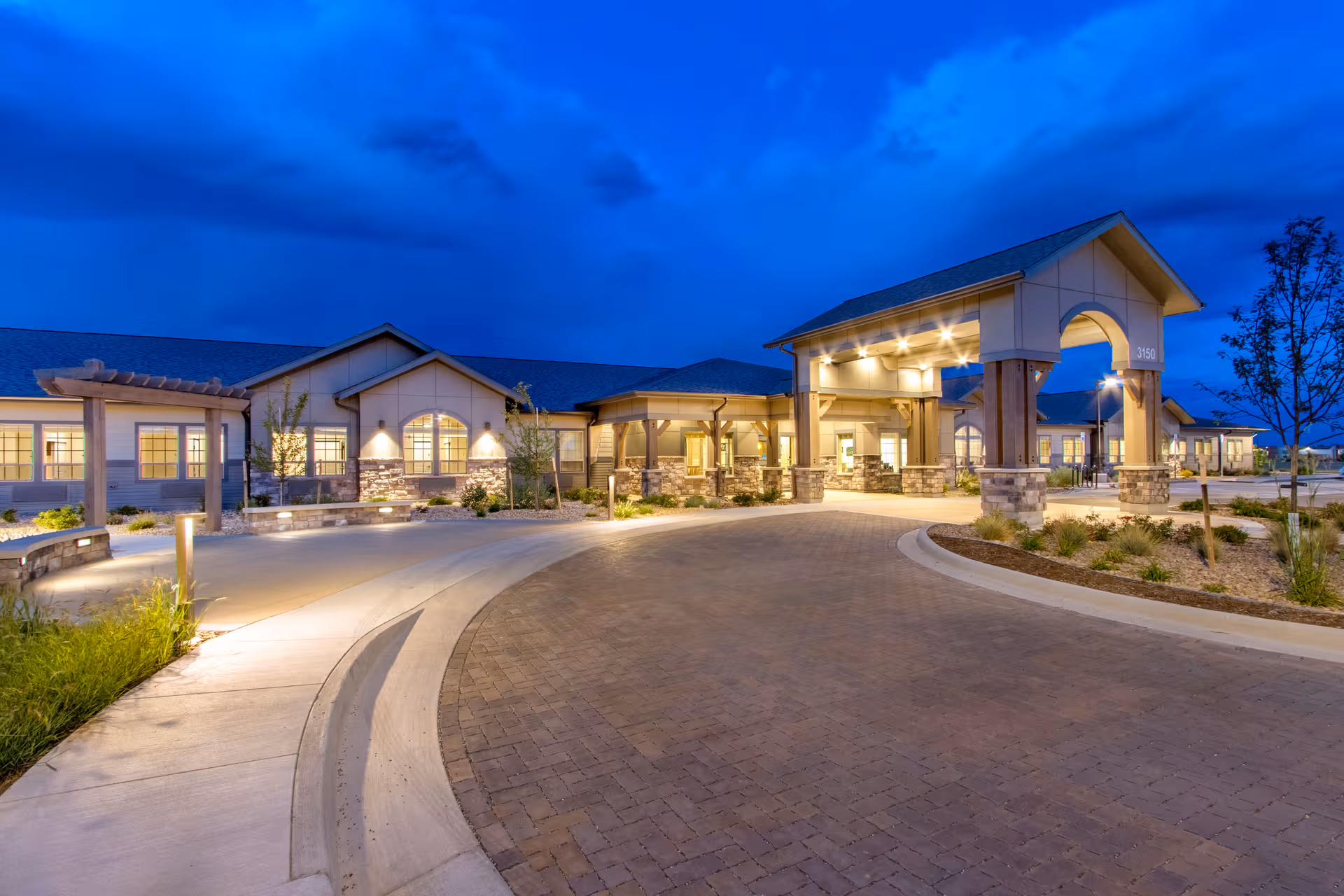 Well-lit senior living facility entrance with curved brick driveway at dusk.