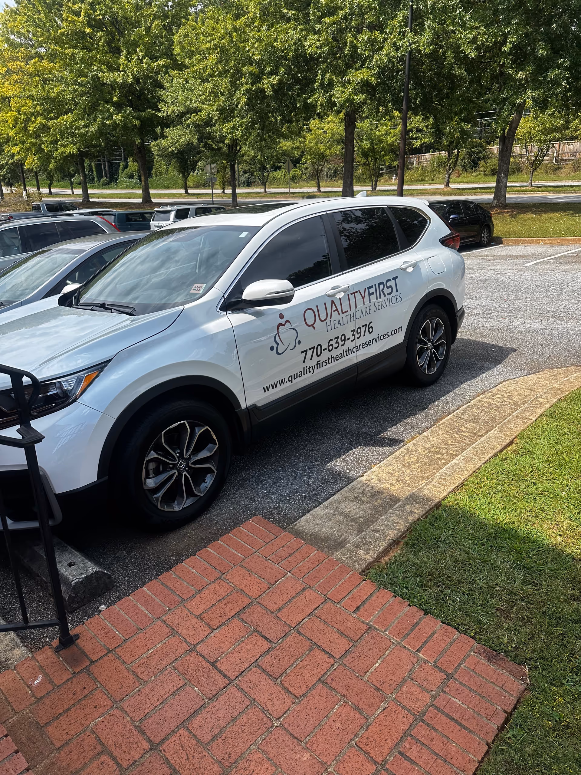 White Quality First Healthcare Services SUV parked in a lot beside trees.