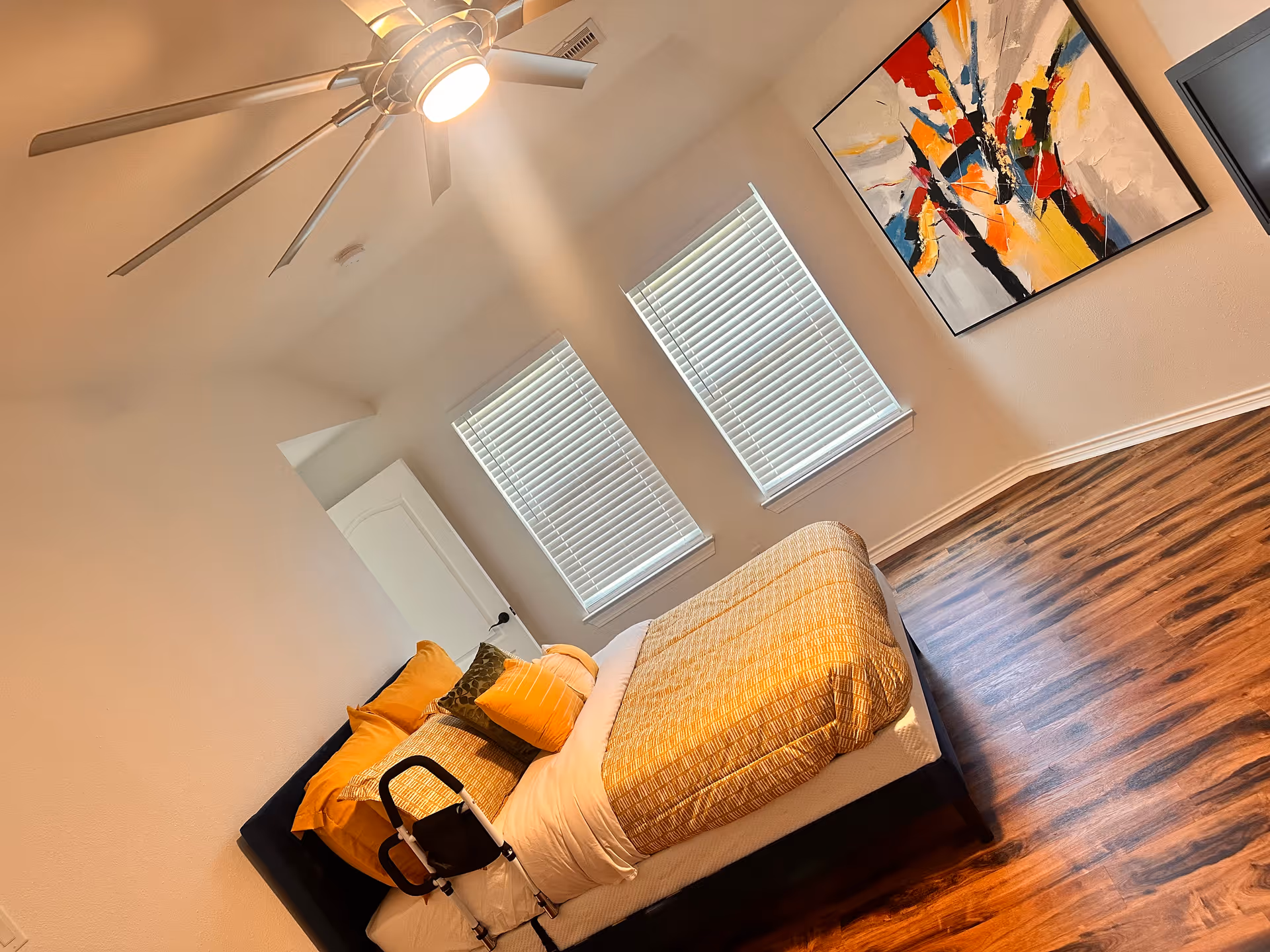 Bedroom with a bed, orange-yellow bedding, abstract wall art, windows, and a ceiling fan.