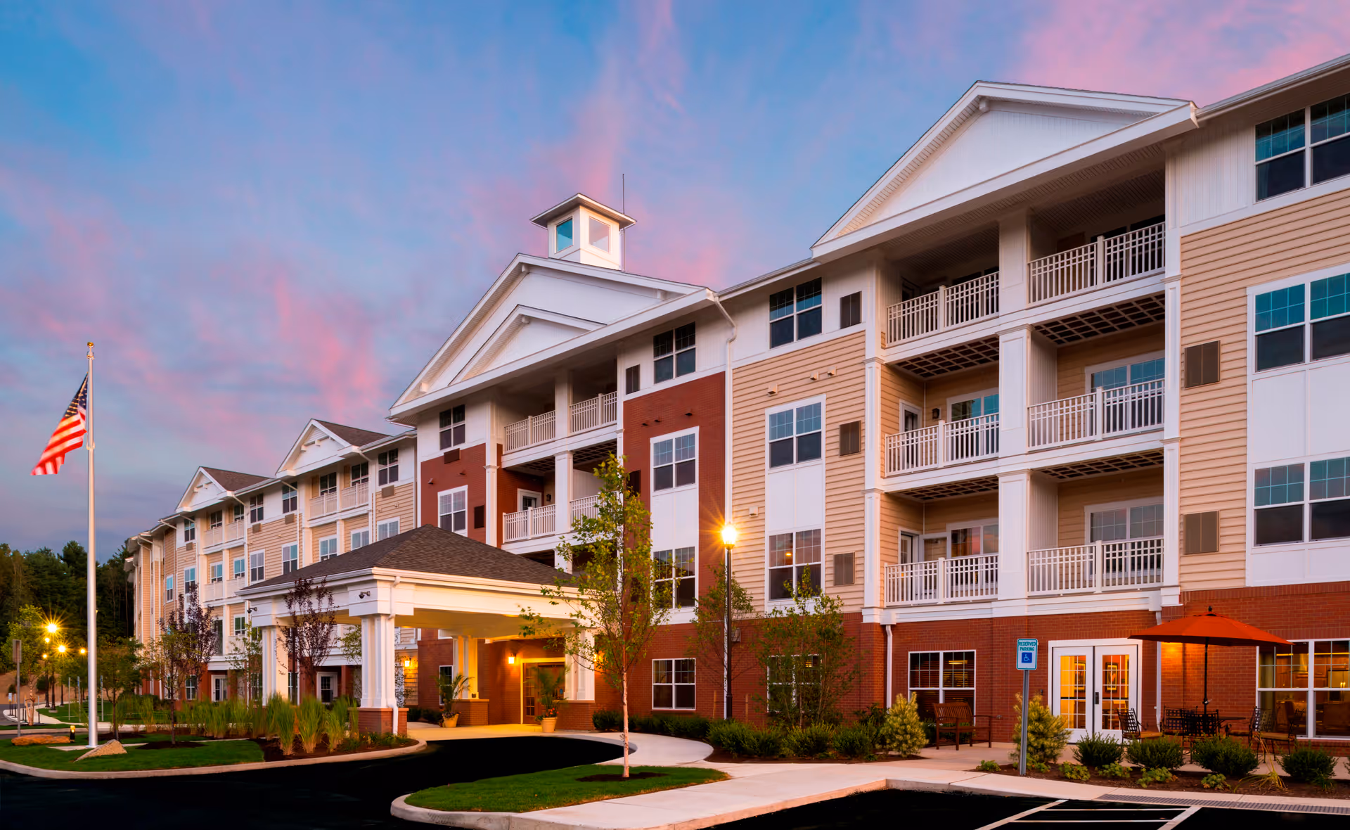 Exterior view of a multi-story senior living facility building at dusk with a covered entrance, balconies, landscaped greenery, an American flag on a flagpole, and a pink and blue sky in the background.