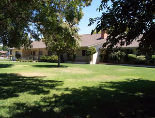 Single-story assisted living building with a covered porch, manicured lawn and shade trees in front.