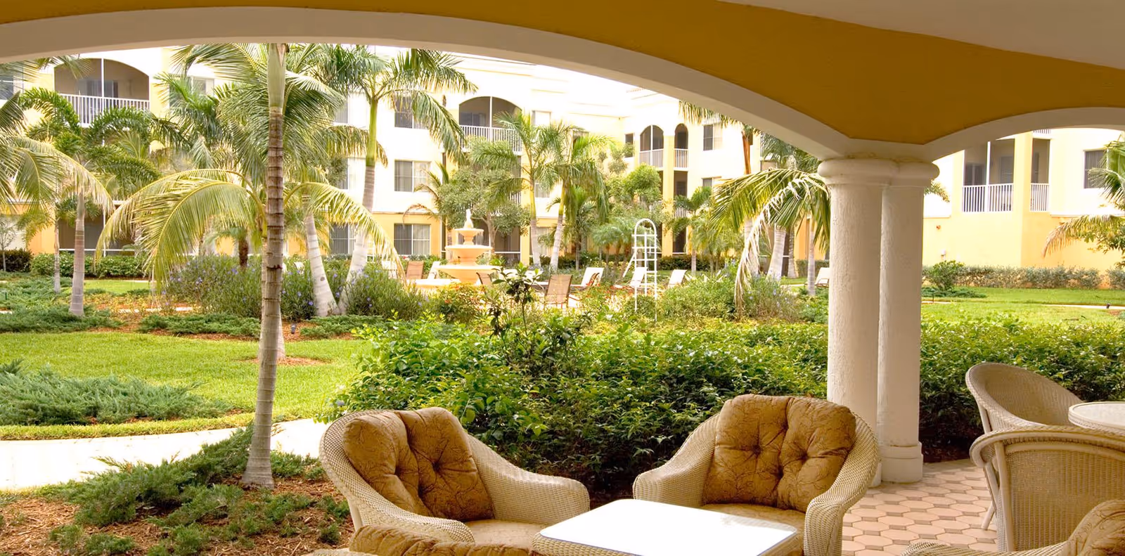Covered patio with wicker chairs and a table overlooking a landscaped courtyard with palm trees and a fountain.