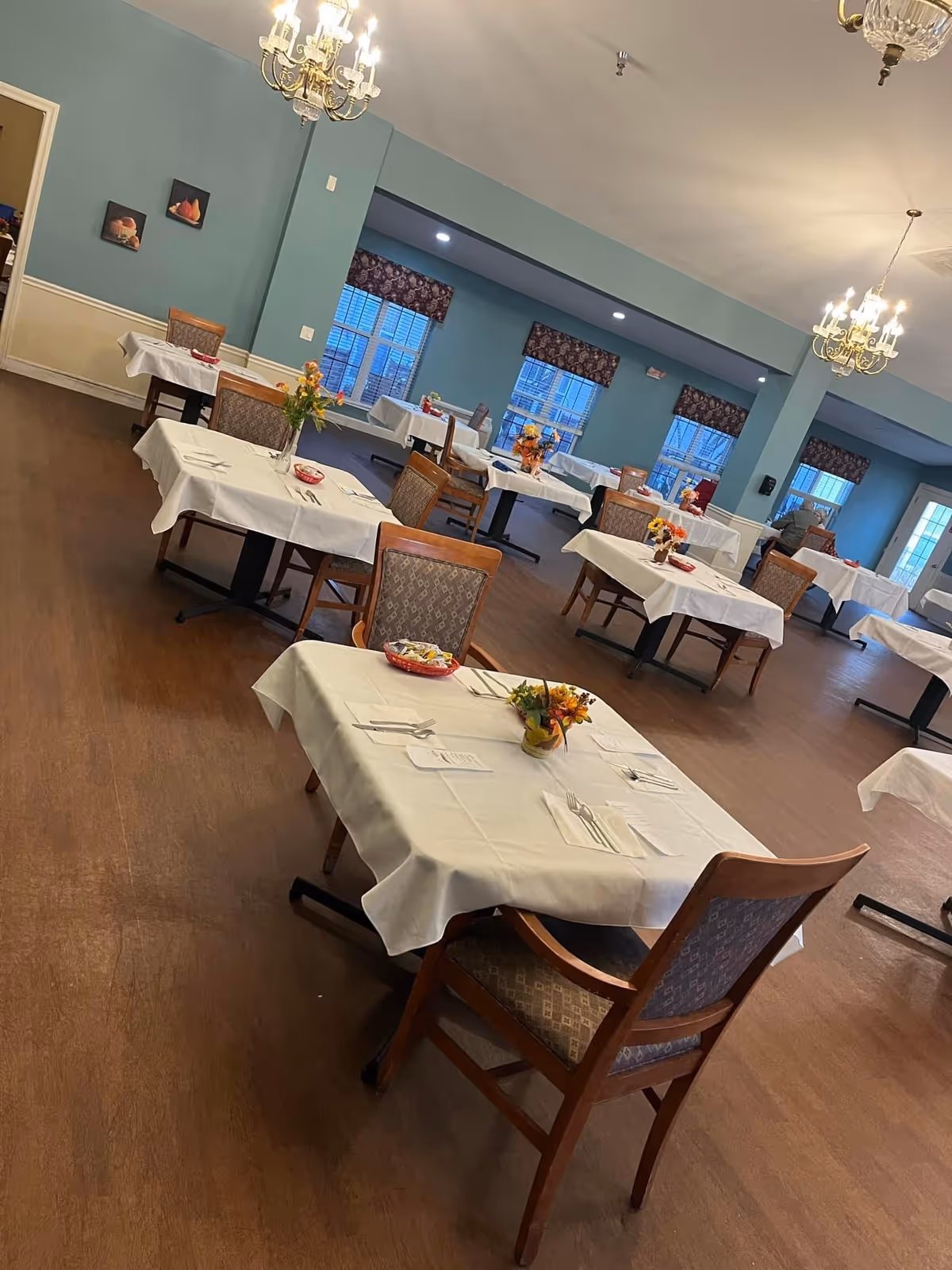 Dining room at Crown Care Center with neatly set tables, chairs, and chandeliers.
