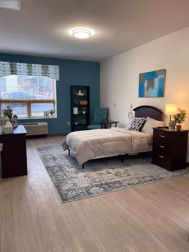 A furnished senior living bedroom featuring a bed, nightstands, rug, and teal accent wall.
