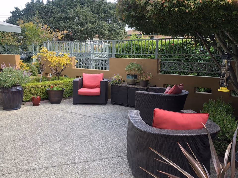 Outdoor patio with wicker seating, red cushions, potted plants, and railing at a senior living facility.