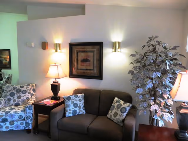 A cozy living room area with a brown loveseat adorned with two floral patterned pillows, a matching floral armchair, a wooden side table with a lamp and magazines, a framed artwork on the wall, two wall-mounted lights, and a large leafy potted plant.