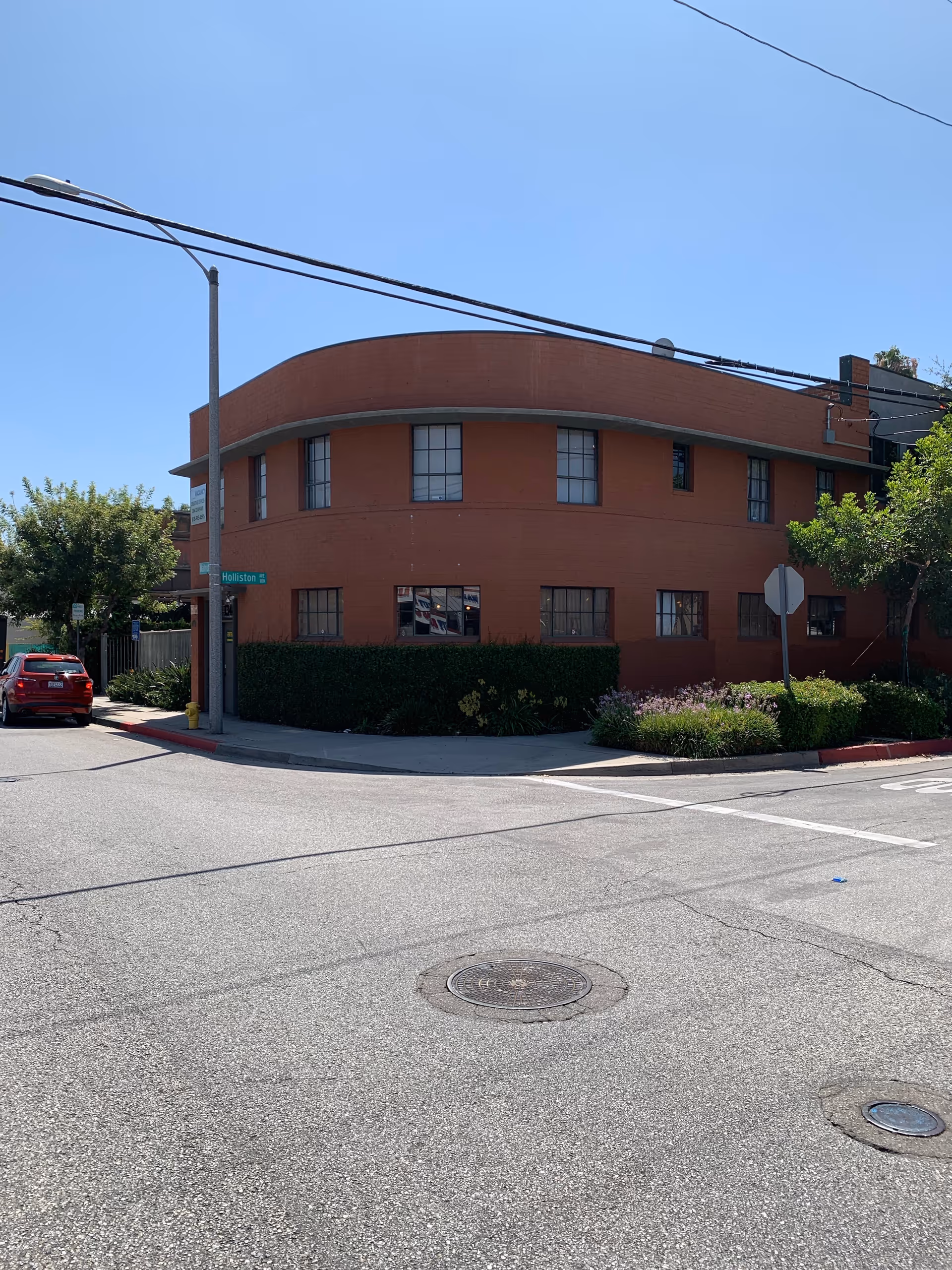 A curved brick building on a street corner with shrubs and a car