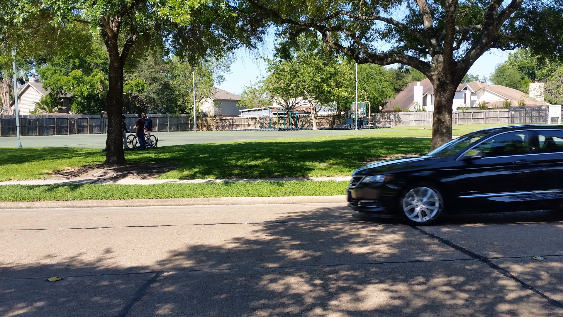 Tree-lined park beside a street, with a passing black car and playground equipment visible.