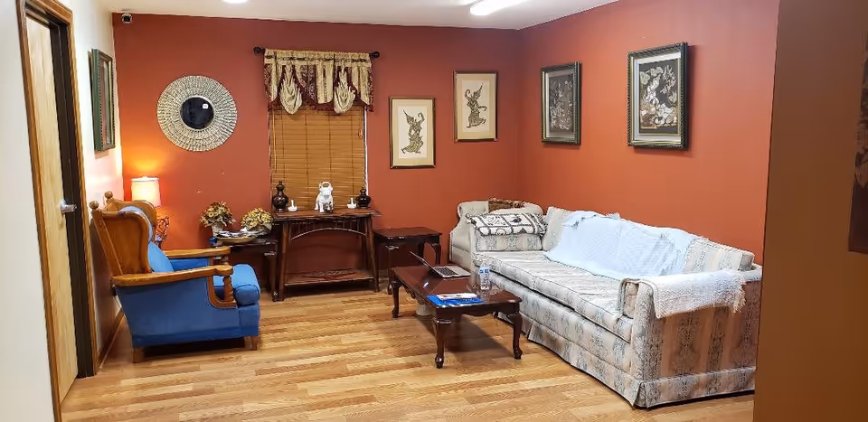 A cozy living room with a patterned sofa, blue armchair, wooden coffee table, red painted walls, framed artwork and hardwood floors.