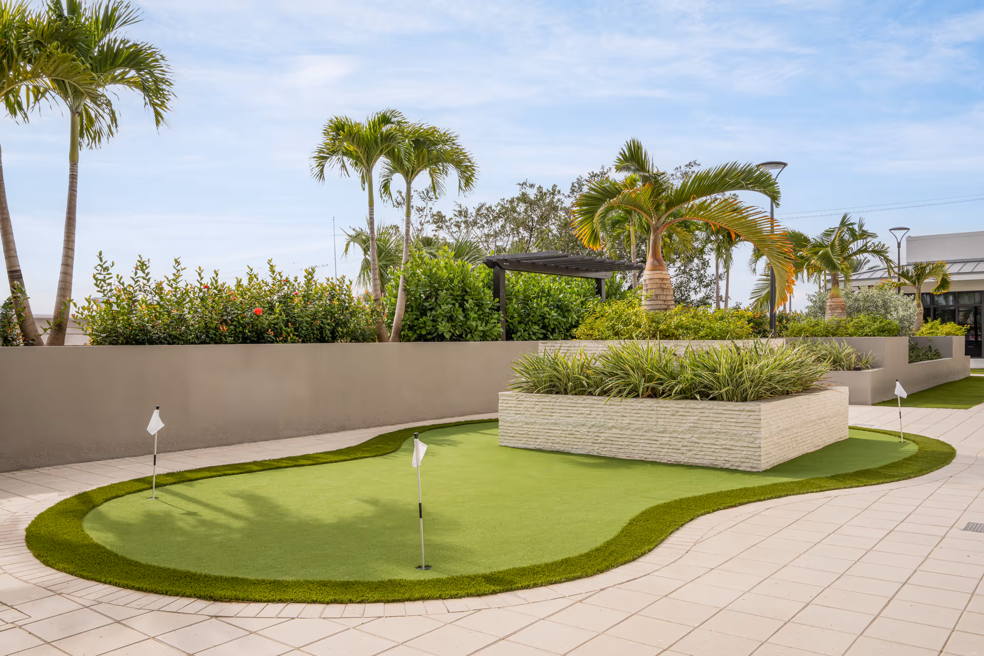 Outdoor putting green with three small flags surrounded by tiled flooring and landscaped with palm trees and bushes under a partly cloudy sky.