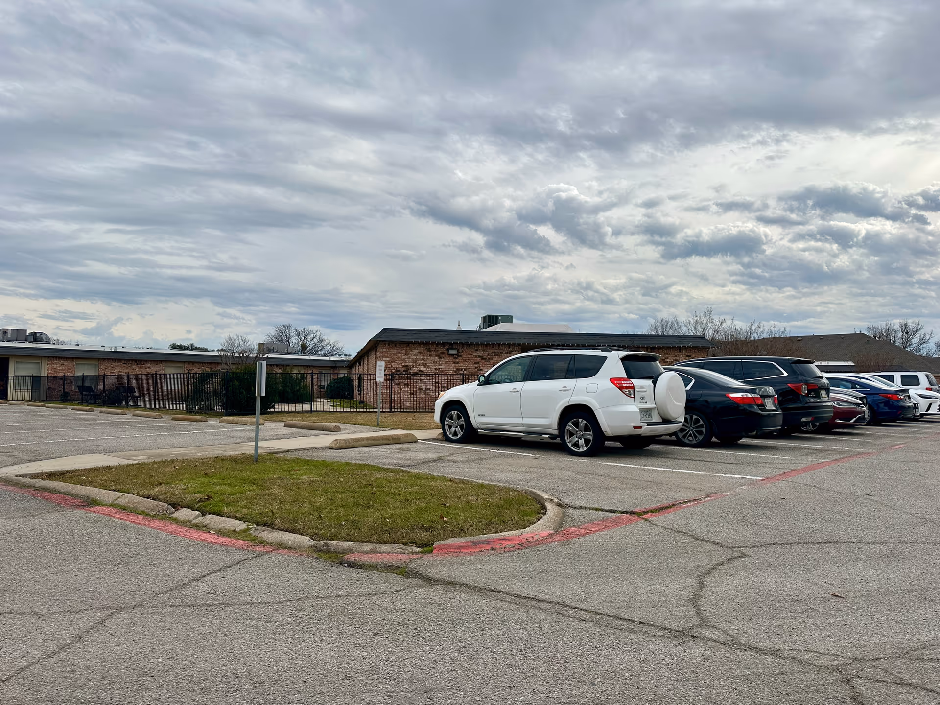 Parking lot with several parked cars in front of a single-story brick senior living facility under a cloudy sky.