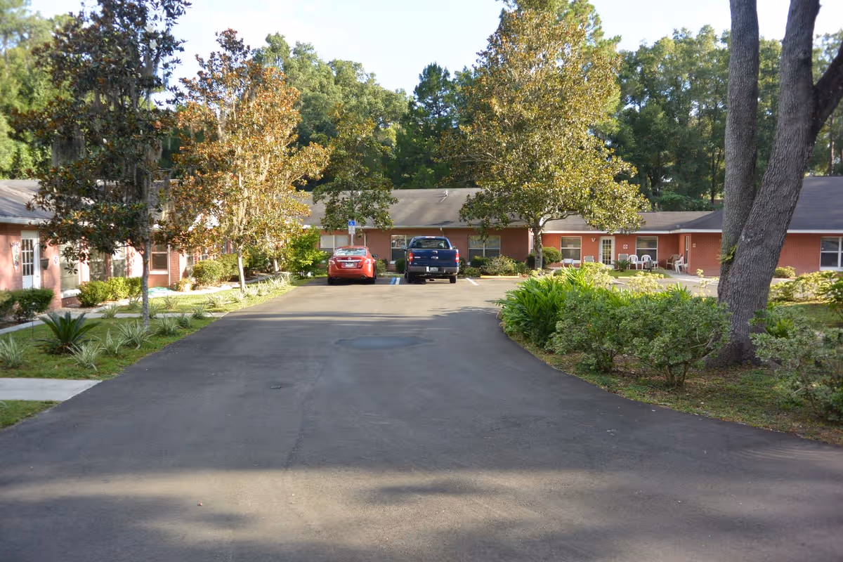 Paved driveway leading to a single-story brick assisted living building with parked cars and landscaped trees.
