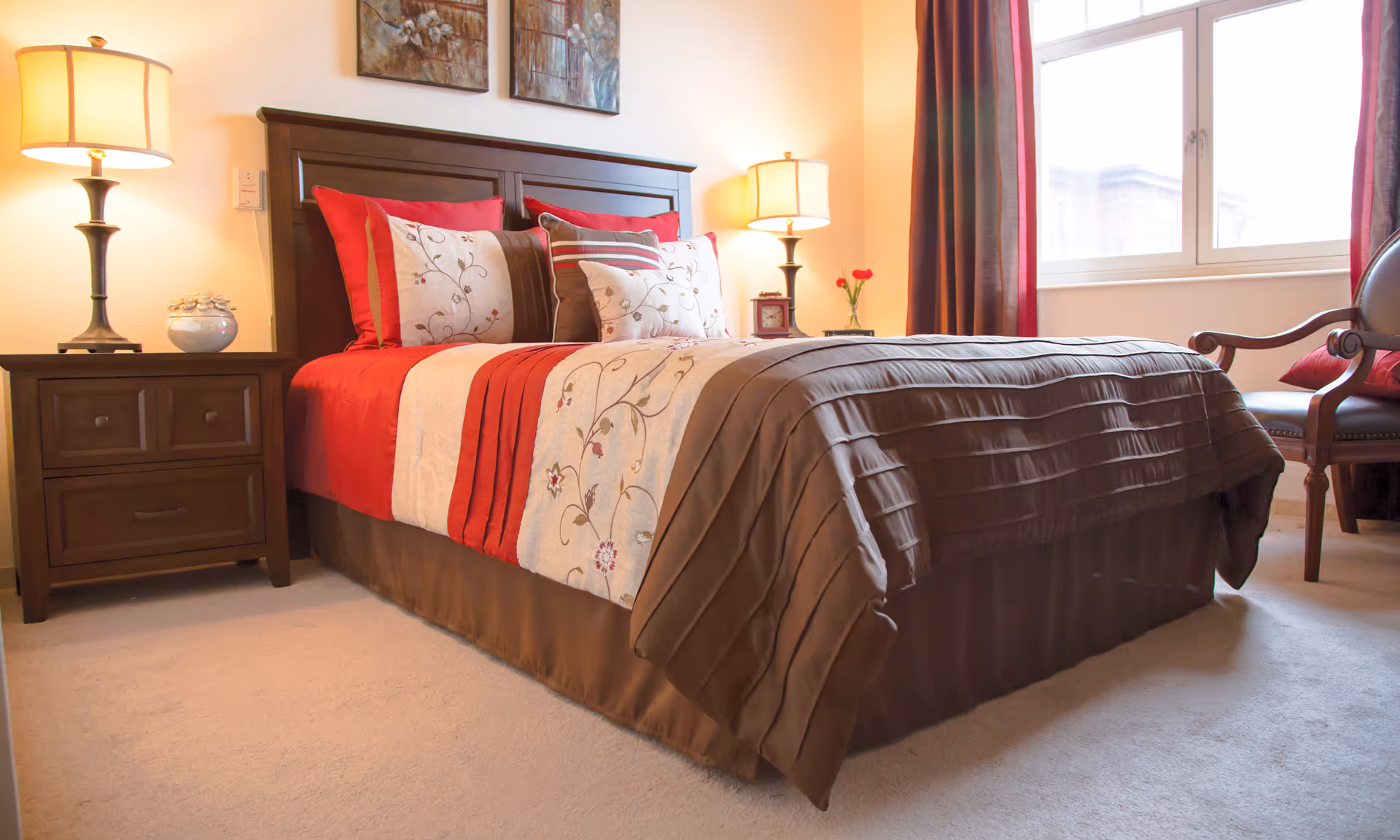 A neatly made bedroom with a dark wood bed, red accents, lamps, and a window