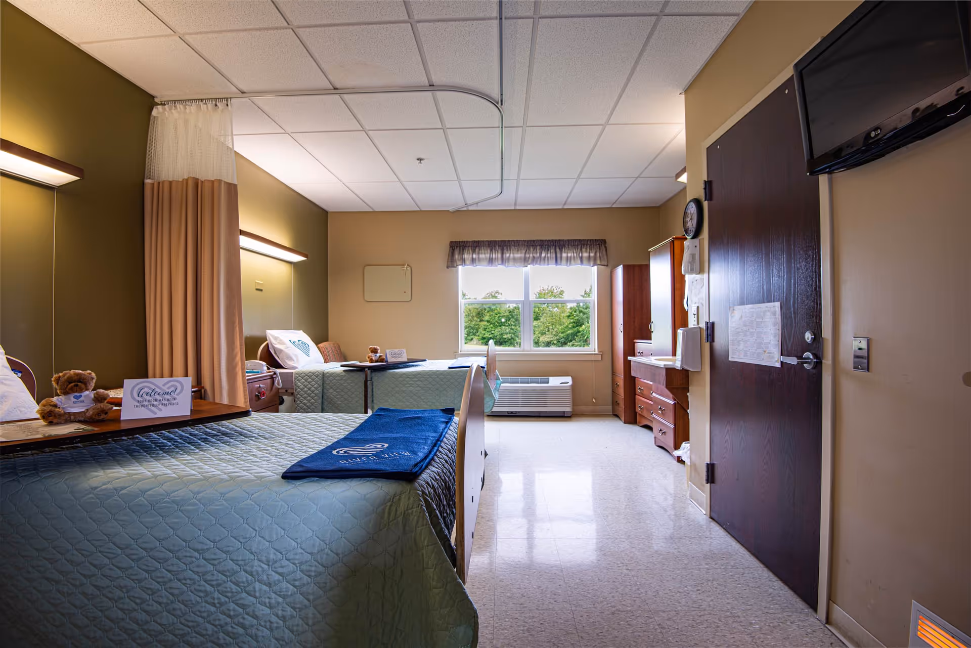 Two hospital-style beds with teal quilts, curtains, and a window view in a patient room.