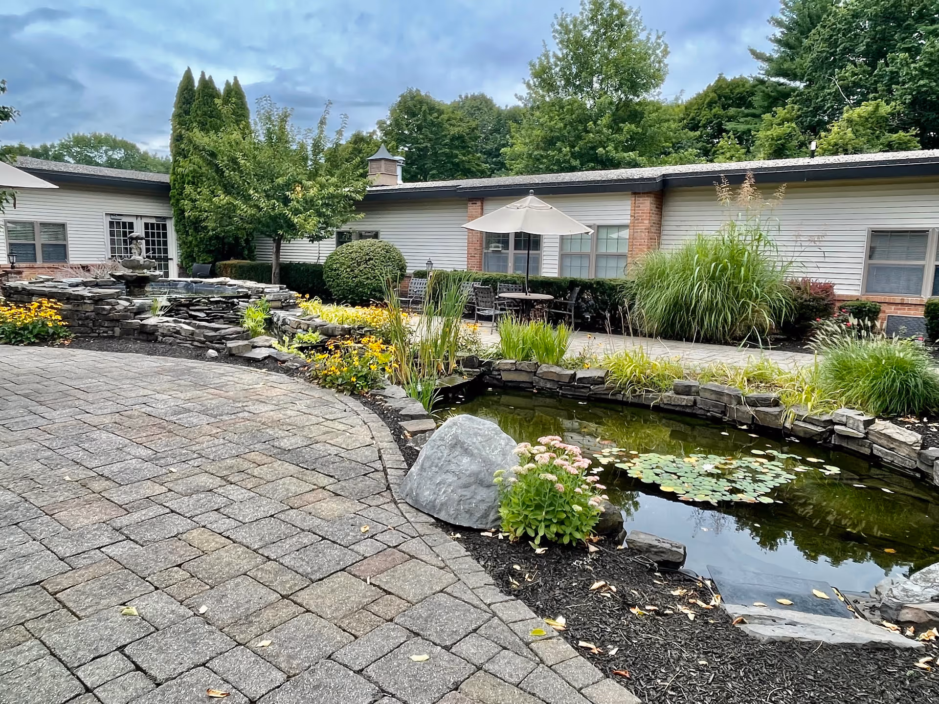 Outdoor garden area at a senior living facility featuring a stone-paved walkway, a small pond with lily pads, a rock, and various plants and flowers. In the background, there is a building with windows, patio tables, chairs, and an umbrella. Trees and shrubs surround the area under a cloudy sky.