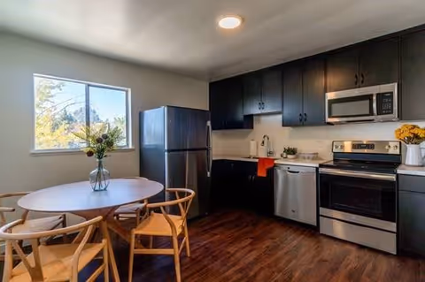 Modern kitchen with dark cabinets, stainless steel appliances, and a round dining table near a window.