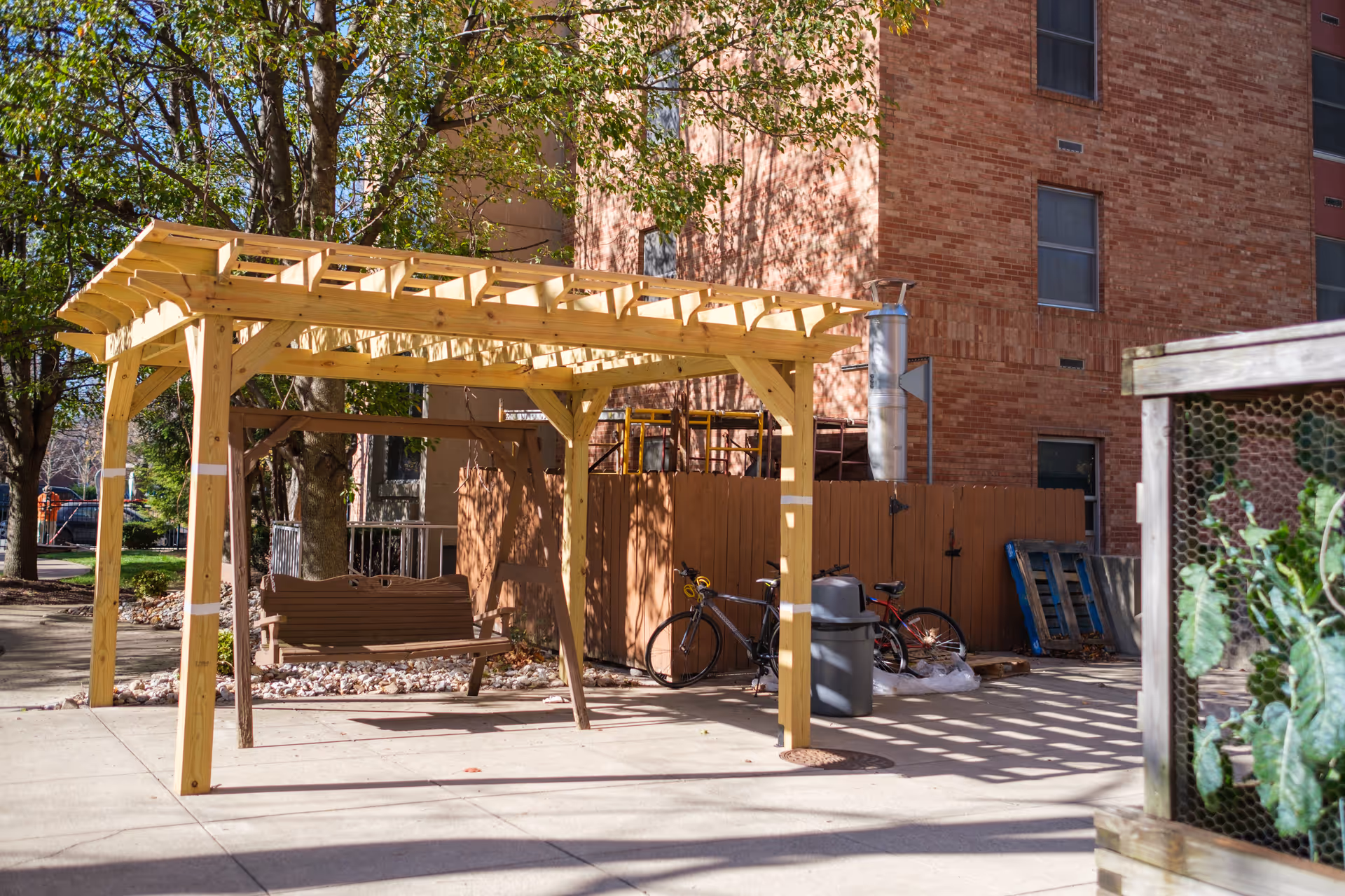 Outdoor area with a wooden pergola structure providing partial shade over a wooden swing bench. Behind the pergola, there are two bicycles leaning against a brown wooden fence. A brick building with several windows is visible in the background, along with some trees and a small garden area with plants enclosed by a wire mesh on the right side.
